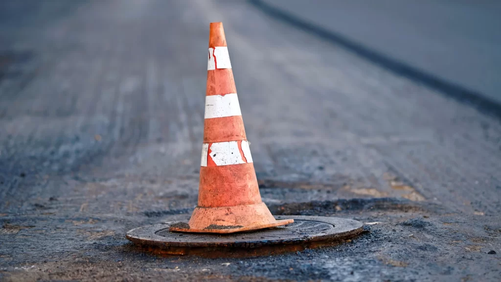 Orange traffic cone stands on manhole. Road repair works, asphalt paving, pylon to mark an obstacle or hole on road. Traffic cone stands on hatch. Road reconstruction, renew old asphalt surface