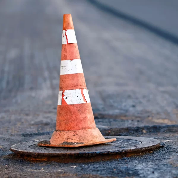 Orange traffic cone stands on manhole. Road repair works, asphalt paving, pylon to mark an obstacle or hole on road. Traffic cone stands on hatch. Road reconstruction, renew old asphalt surface