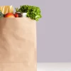 Various grocery items in paper bag on white table opposite gray wall. Bag of food with fresh vegetables, fruits, pasta and canned goods. Food delivery, shopping or donation concept. Copy space.