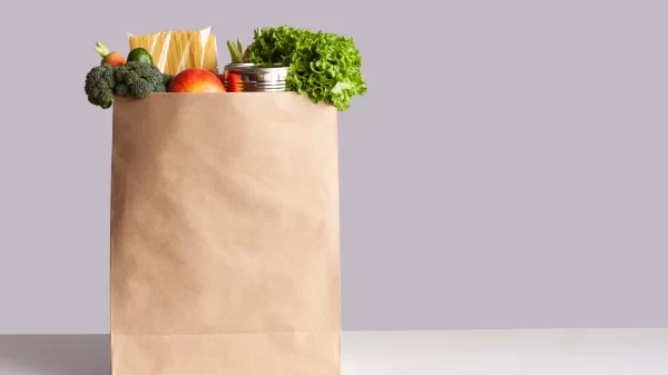 Various grocery items in paper bag on white table opposite gray wall. Bag of food with fresh vegetables, fruits, pasta and canned goods. Food delivery, shopping or donation concept. Copy space.