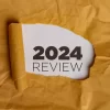 A yellow piece of paper with a hole torn in it. Through the hole, a white piece of paper is visible with the text "2024 REVIEW" written in black. This represents the concept of reviewing the year 2024