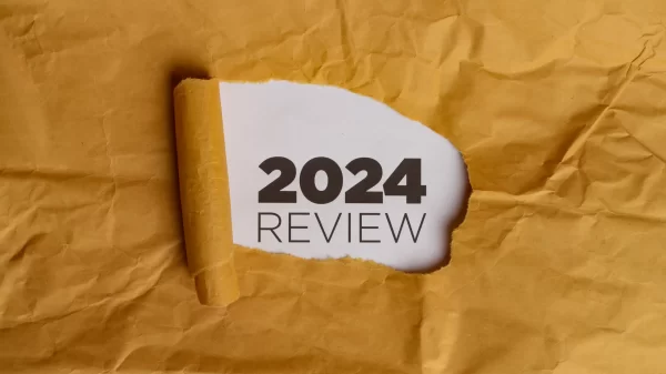 A yellow piece of paper with a hole torn in it. Through the hole, a white piece of paper is visible with the text "2024 REVIEW" written in black. This represents the concept of reviewing the year 2024