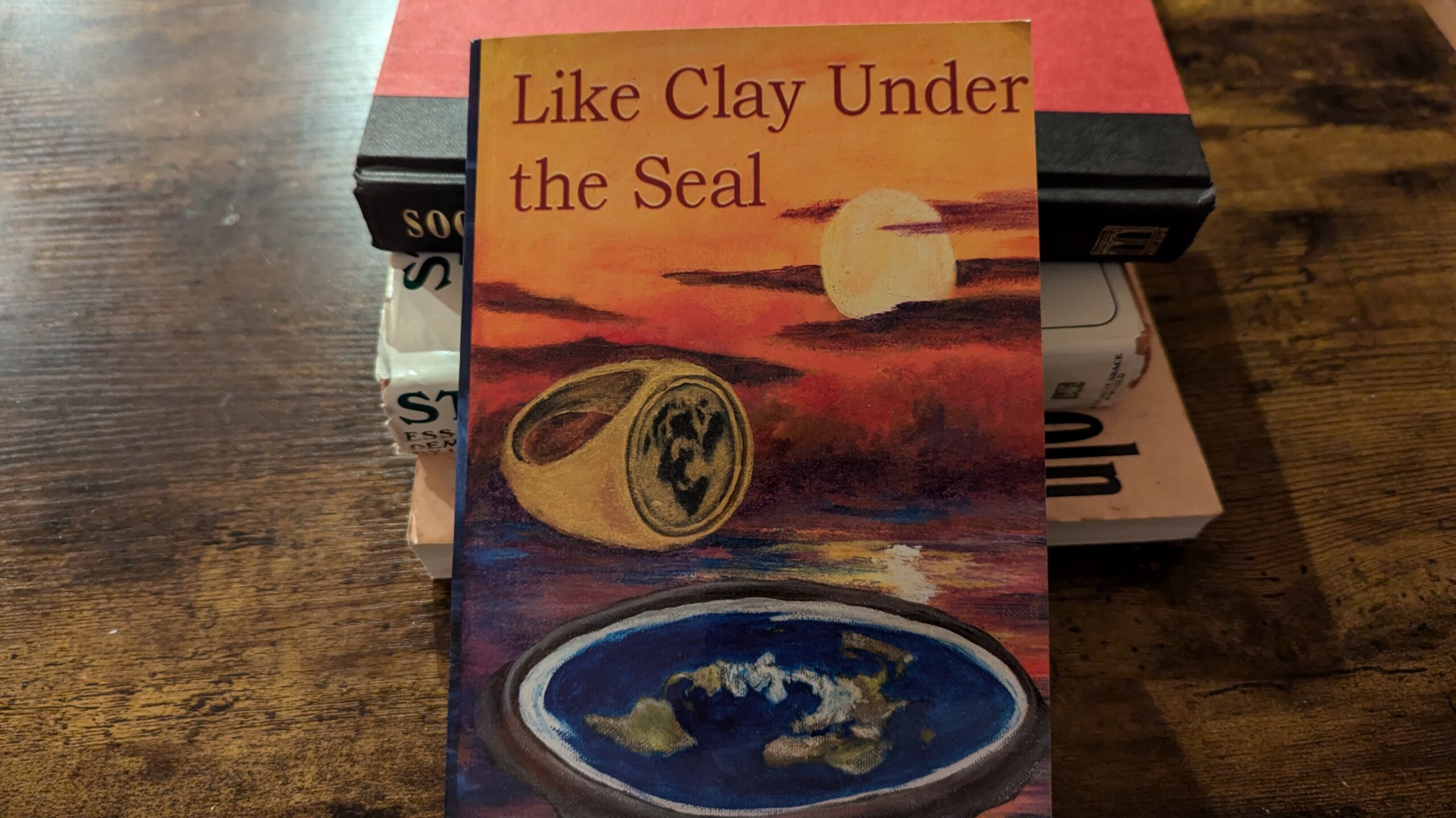 A photo of “Like Clay Under the Sun” resting against a pile of other books.